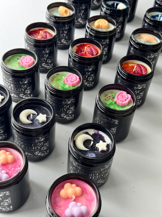A range of Court of Mystery candles with lids off, showing the variations for each court with different colours, glitter and wax shapes on top.
