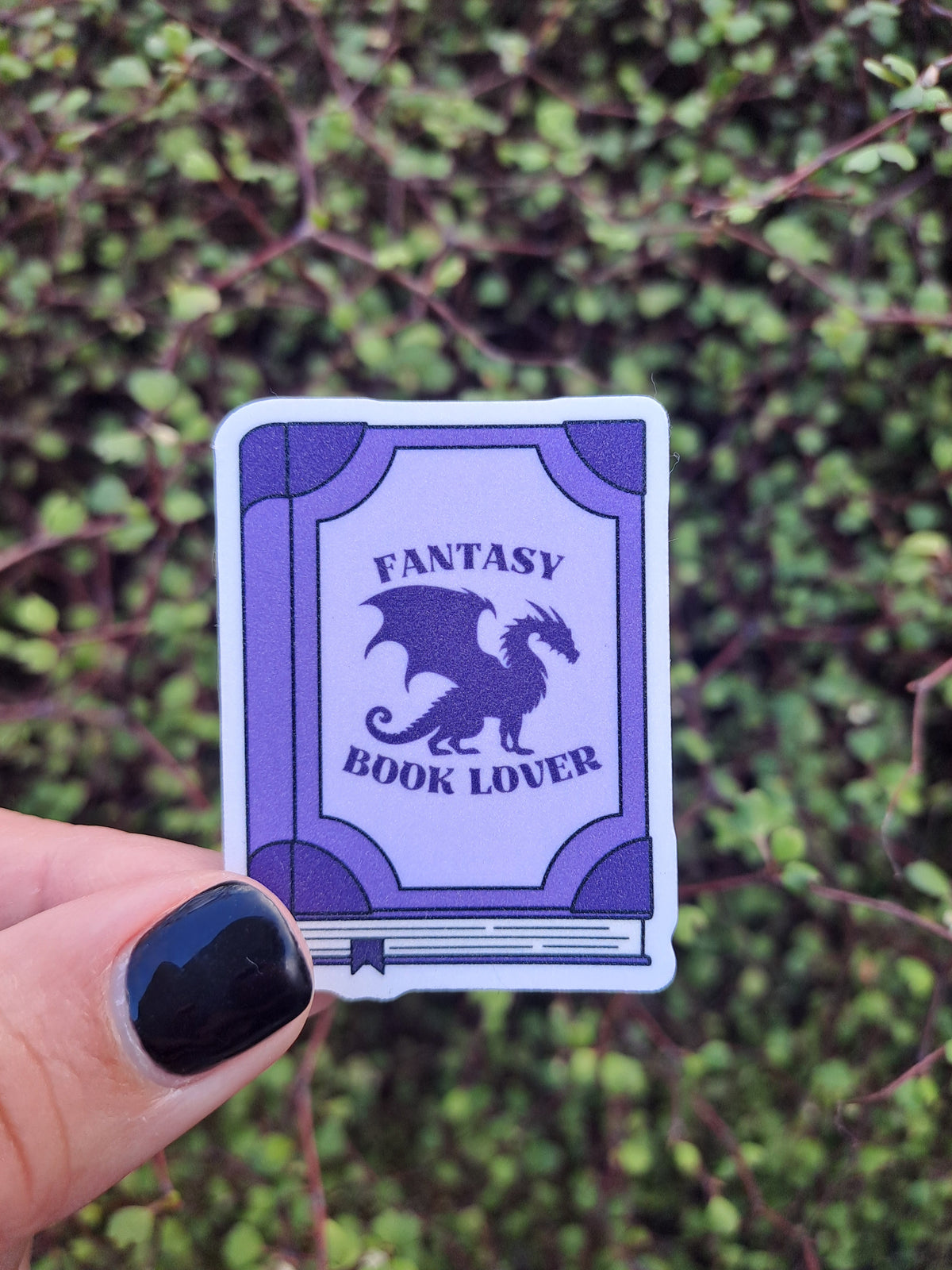 Purple book sticker with a dragon and text: Fantasy Book Lover. Held by Steph outside, the sticker is glittery.